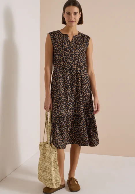 Midikleid mit Split Neck und Print creamy mocha Frau trägt ärmelloses, knielanges Leopardenmuster-Kleid mit Strohtasche und braunen Loafers vor beige Hintergrund.