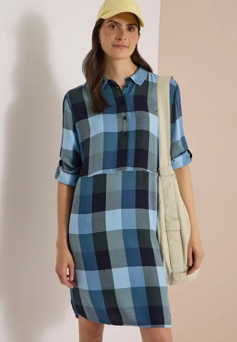 Femme en robe-chemise à carreaux bleus, casquette et sac beige, devant un fond neutre.