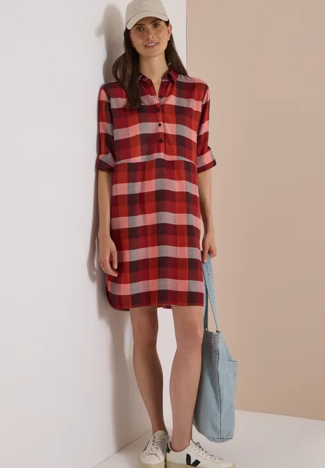 Kniehoge jurk met ruitjespatroon raw chocolate brown Eine Frau trägt ein rotes kariertes Hemdkleid, weiße Sneakers, Basecap und eine blaue Tasche.