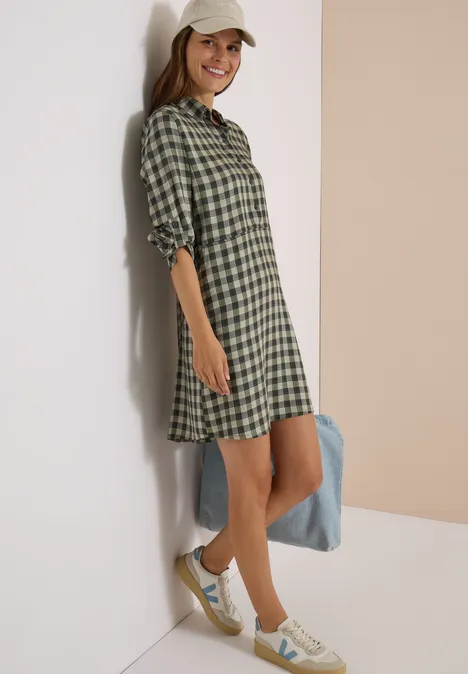 Robe arrivant aux genoux, à petits carreaux khaki Femme appuyée contre un mur, en robe à carreaux et casquette, baskets blanches aux pieds, sac bleu clair à la main.