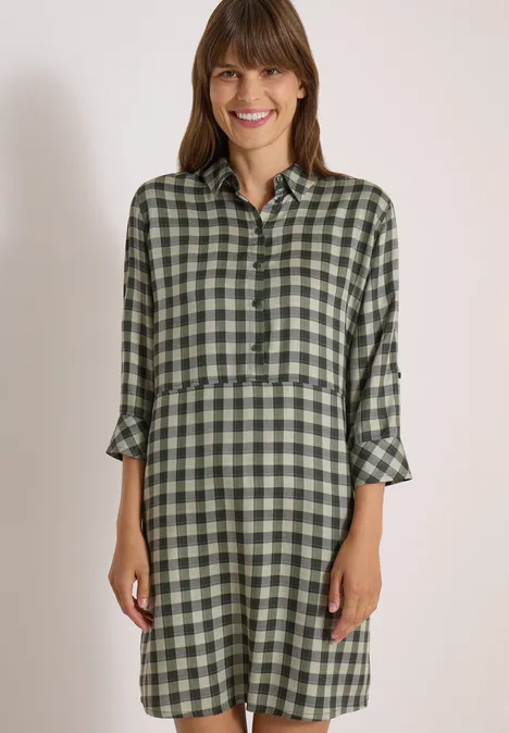 Robe arrivant aux genoux, à petits carreaux khaki Femme en robe chemise à carreaux verts, souriante, posant devant un fond uni.