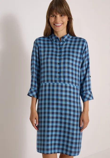 Eine lächelnde Frau trägt ein blau kariertes Hemdkleid mit 3/4-Ärmeln und Kragen.