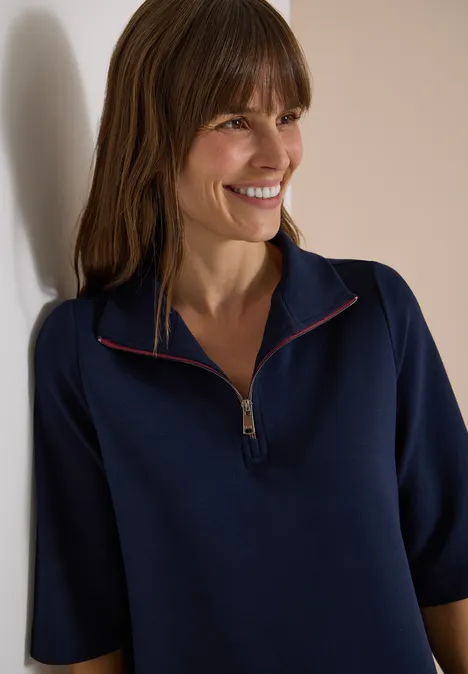 Eine Frau lehnt lächelnd an einer Wand und trägt einen marineblauen Pullover mit kurzem Reißverschluss.