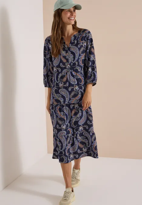 Frau in blau-weiß-rotem Paisley-Midikleid mit 3/4-Ärmeln und Baseballcap vor hellem Hintergrund.