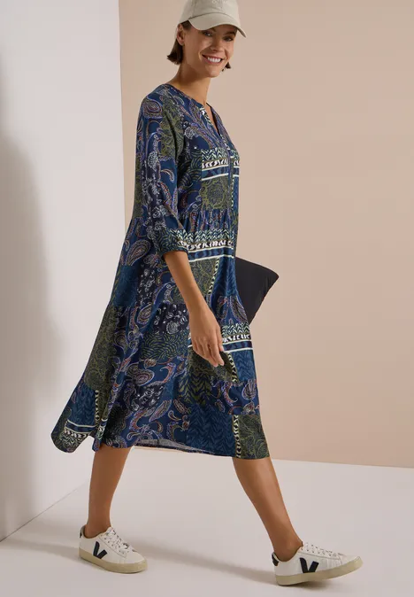 Frau in blau gemustertem Kleid und weißen Sneakers, geht lächelnd vor hellem Hintergrund.