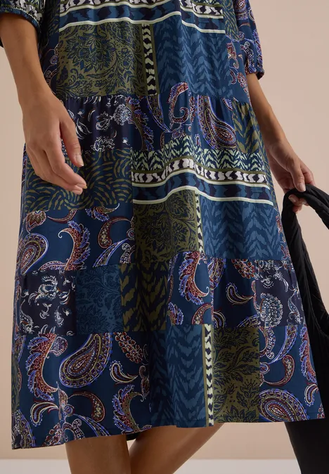 Knielanges Patchwork-Kleid mit Paisley- und Blumenmuster in Blau- und Grüntönen, schwarze Tasche in der Hand.
