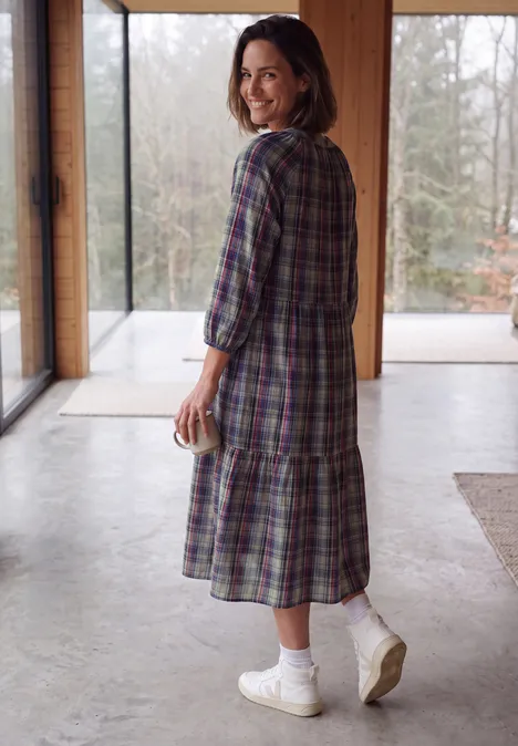 Vrouw in geruite jurk, witte sokken en sneakers, houdt een mok vast en lacht bij het raam.