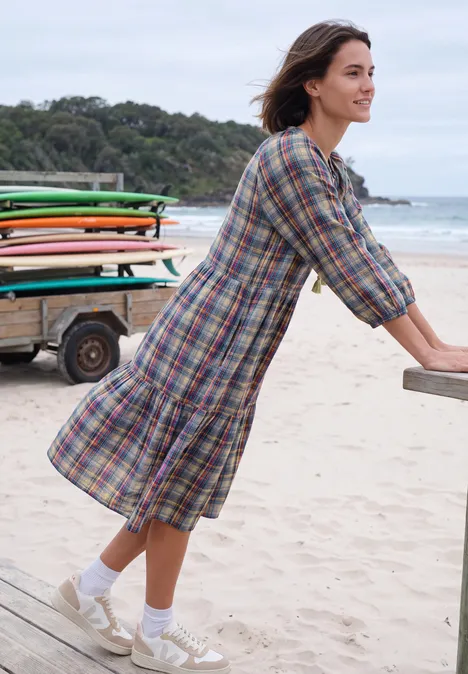 Knielanges Kleid mit Split Neck und Print creamy mocha Eine Frau lehnt am Strandgeländer, trägt ein kariertes Kleid, Socken und Sneakers, im Hintergrund Surfbretter.