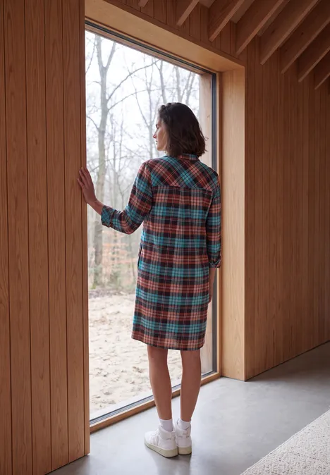 Knielanges Flanellkleid mit Print dark russet red Frau mit kariertem Mantel steht in einem Holzbau an einem hohen Fenster und schaut nach draußen.