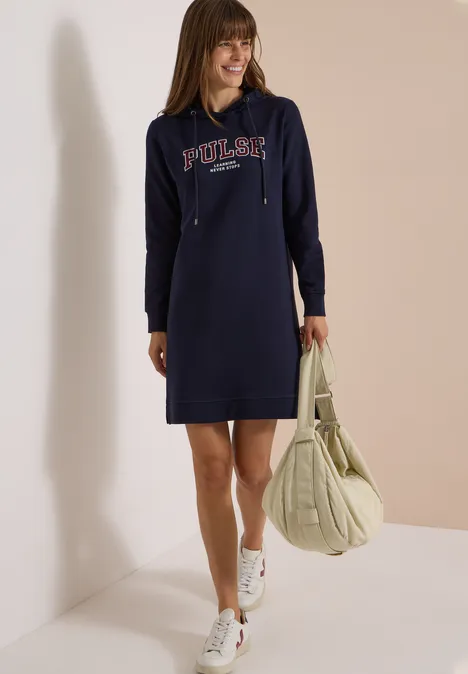 Frau in blauem Hoodie-Kleid mit Aufschrift, weiße Sneaker, großer Beutel, stehend vor heller Wand.