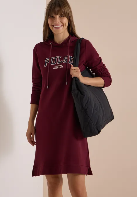 Frau in dunkelrotem Kapuzenkleid mit Schriftzug 'Pulse', lächelnd und trägt eine große schwarze Tasche.