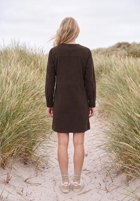 Une femme en robe marron, de dos, se tient sur des dunes de sable entourées d’herbes de plage.