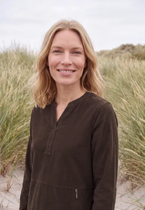 Femme blonde souriante debout dans des dunes, vêtue d’un haut sombre.