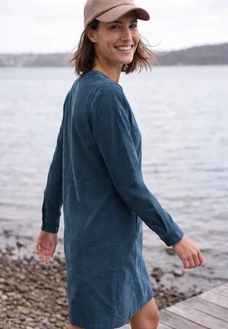 Eine lächelnde Frau in einem blauen Jeanskleid und Baseballcap steht am Ufer und blickt zurück.