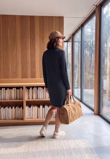 Kniehoge jurk met col urban dark blue Een vrouw in jurk en pet met reistasje in een lichte kamer met boekenkast en grote ramen.