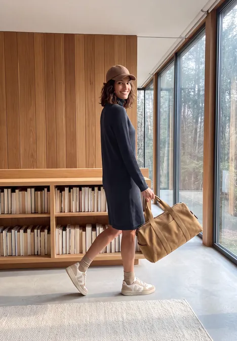Kniehoge jurk met col urban dark blue Vrouw in blauwe jurk en pet met tas in een moderne ruimte met houten wand en grote ramen.