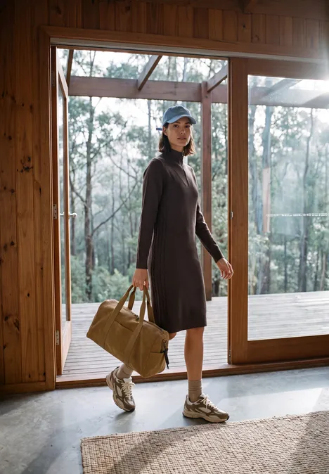 Vrouw in gebreide jurk en sneakers met weekendtas voor grote ramen met uitzicht op het bos.