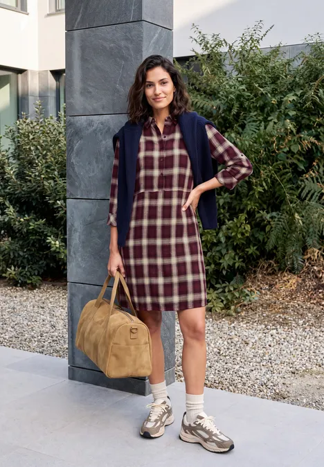 Frau im karierten Kleid mit Jacke um die Schultern, Sneakers und einer beigen Tasche vor Haus und Pflanzen.