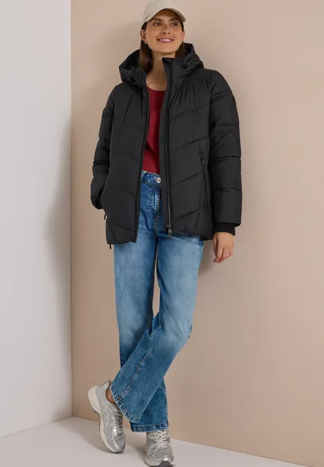 Frau in schwarzer Steppjacke, rotem Pullover, blauen Jeans, silbernen Sneakern und Cap vor beigem Hintergrund.