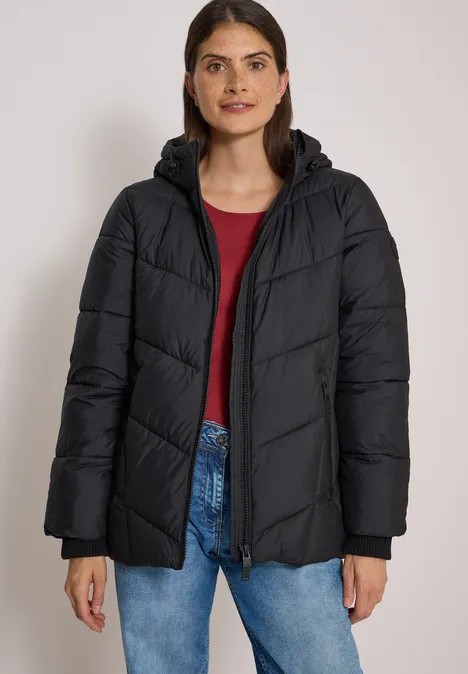 Eine Frau in einer schwarzen Steppjacke mit Kapuze, rotem Oberteil und Jeans vor hellem Hintergrund.