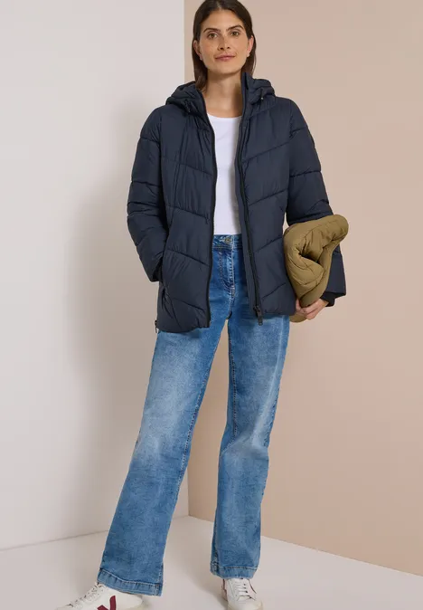 Frau in dunkelblauer Steppjacke, weißem Shirt, blauen Jeans, hält eine olivgrüne Tasche vor neutralem Hintergrund.
