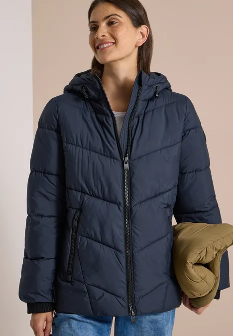Frau trägt dunkelblaue Steppjacke mit Kapuze, lächelt und hält eine gefaltete khakifarbene Jacke.