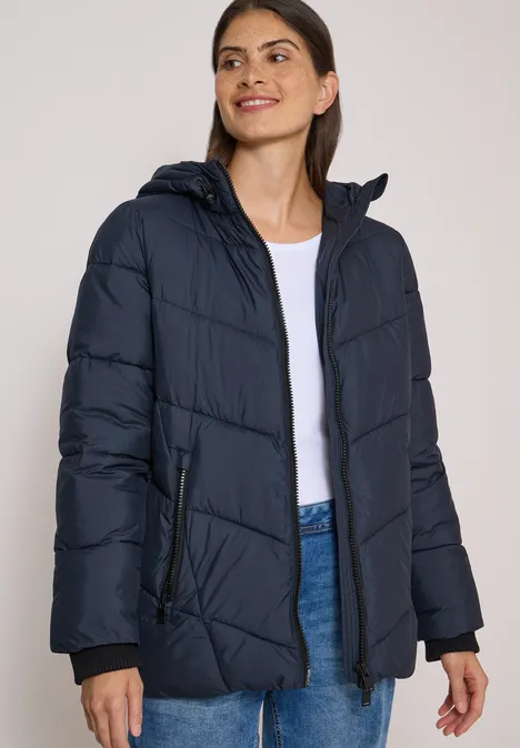 Frau in dunkelblauer Steppjacke, weißem Shirt und Jeans.