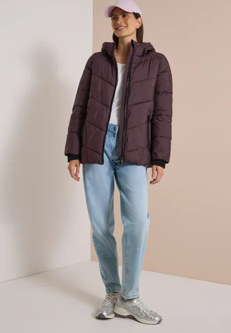 Frau in lila Steppjacke, hellem Shirt, weitem Jeans, Sneakern und rosa Cap vor neutralem Hintergrund.