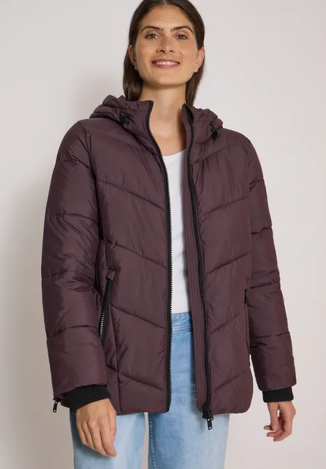 Frau in violettem, gepolstertem Winterparka mit Kapuze lächelt freundlich.