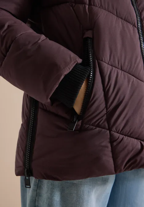 Nahaufnahme einer Person mit dunkelroter Steppjacke, Hand in Tasche, schwarzer Reißverschluss, blaue Jeans.
