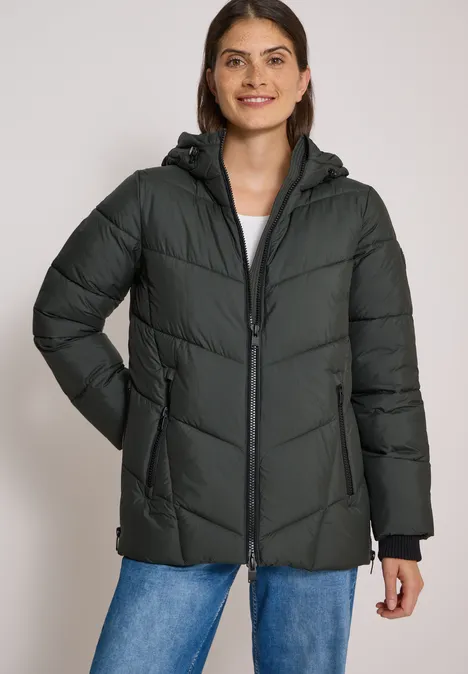 Puffer-Jacke in Midi-Länge mit Kapuze olive night Frau trägt dunkelgrüne, gesteppte Winterjacke mit Kapuze und Reißverschluss, kombiniert mit Jeans.