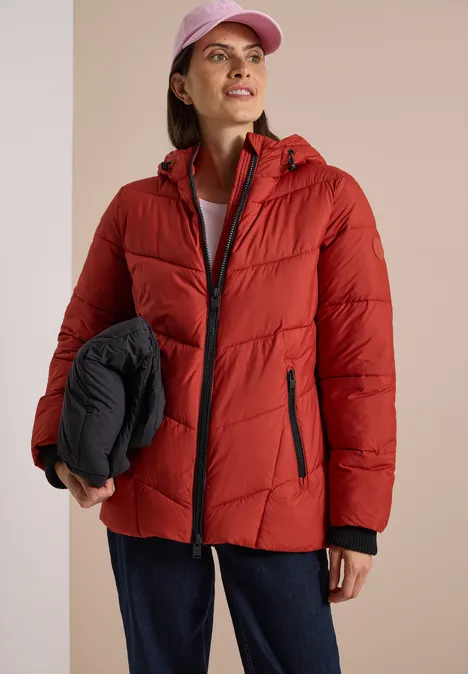 Puffer-Jacke in Midi-Länge mit Kapuze light russet red Frau in roter Steppjacke mit Kapuze, trägt rosa Cap und hält schwarze, zusammengefaltete Jacke, lächelt.