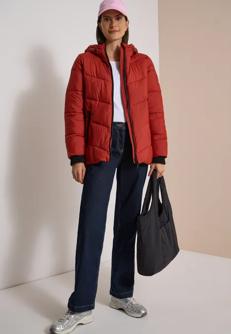 Puffer-Jacke in Midi-Länge mit Kapuze light russet red Frau in roter Steppjacke, weißem Shirt, blauen Jeans und Sneakern trägt pinke Kappe und schwarze Tasche.