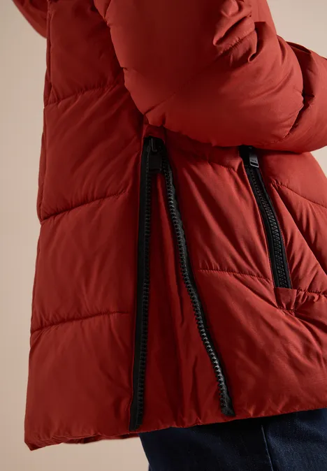 Puffer-Jacke in Midi-Länge mit Kapuze light russet red Roter Steppmantel mit schwarzem Reißverschluss und seitlichen Zipper-Details, getragen zu blauer Hose.