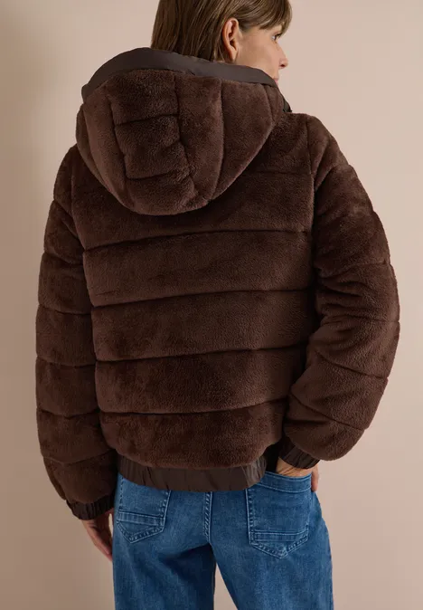 Person von hinten in brauner, gepolsterter Kapuzenjacke und blauen Jeans vor neutralem Hintergrund.