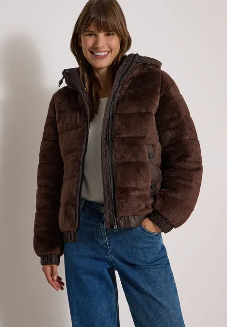 Junge Frau in brauner Steppjacke und blauen Jeans lächelt vor neutralem Hintergrund.