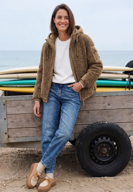 Eine lächelnde Frau am Strand in Jeans, weißem Shirt und brauner Teddy-Jacke lehnt an einem Bretterwagen.