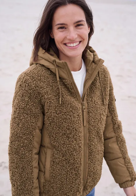 Frau lächelt am Strand in brauner Teddy-Fleece-Jacke mit Kapuze.