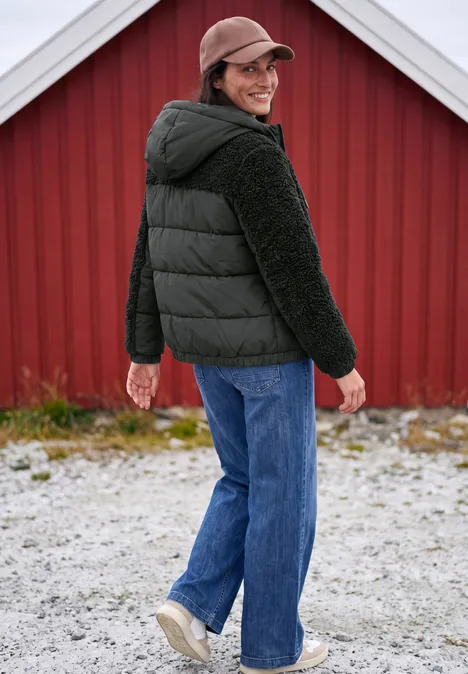Ein lächelnder Mann in schwarzer Steppjacke und Cap blickt zurück vor einem roten Holzhaus, trägt Jeans und Sneaker.