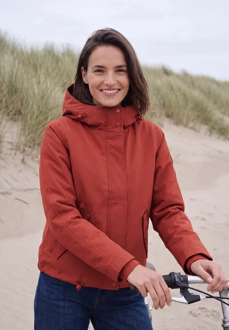 Frau in roter Jacke am Sandstrand, lächelnd, hält Fahrradlenker, Dünen im Hintergrund.
