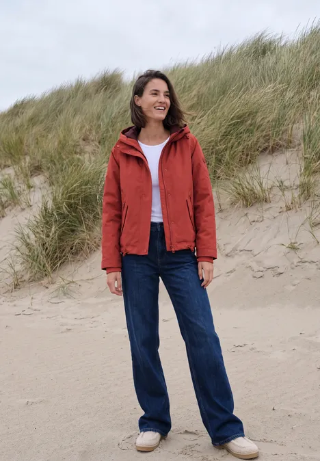 Eine lächelnde Frau in roter Jacke, weißem Shirt und Jeans steht am Strand vor Dünen