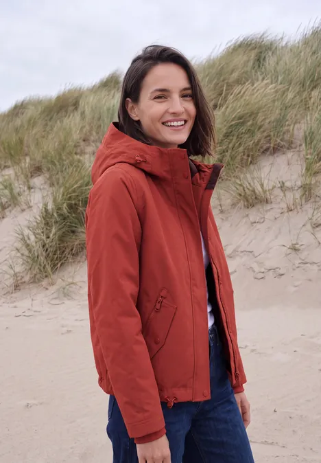 Frau trägt rote Jacke am windigen Dünenstrand und lächelt in Richtung Kamera.