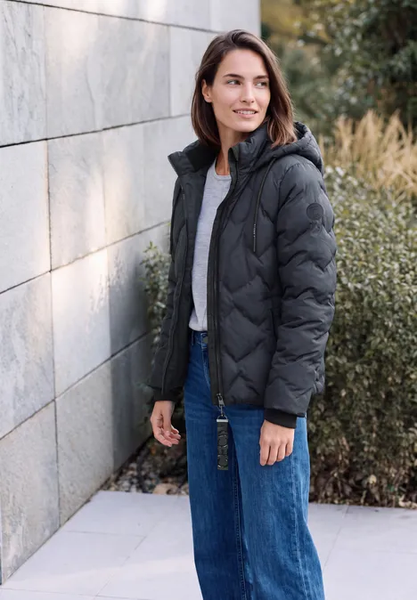 Frau in schwarzer Steppjacke und blauen Jeans steht draußen vor Mauer und Büschen und lächelt.