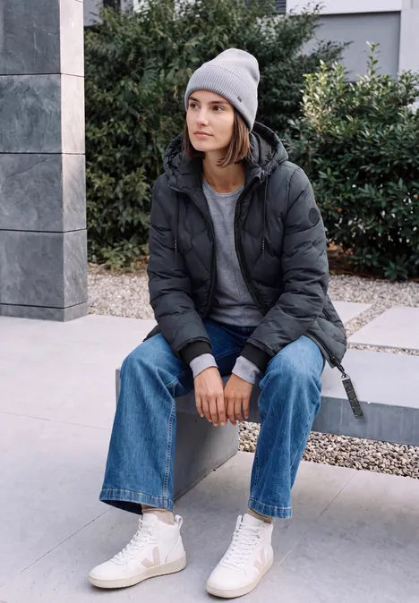 Junge Frau sitzt draußen auf einer Betonbank, trägt Beanie, Daunenjacke, Jeans und helle Sneakers.