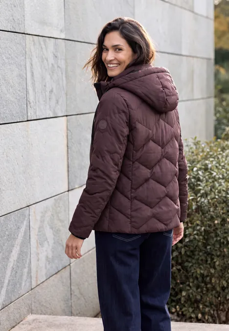 Veste matelassée à capuche et fermeture éclair berry red Frau in dunkelroter Steppjacke mit Kapuze steht draußen an einer Mauer und schaut lächelnd zurück.