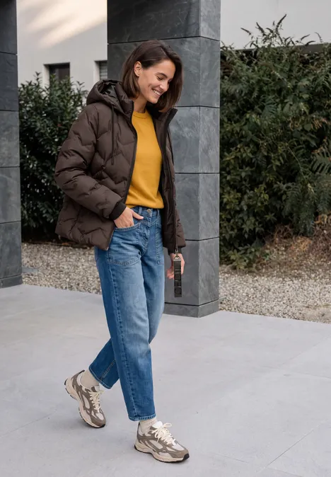 Frau in brauner Steppjacke, gelbem Pullover, Jeans und Sneakern spaziert draußen mit Kaffee