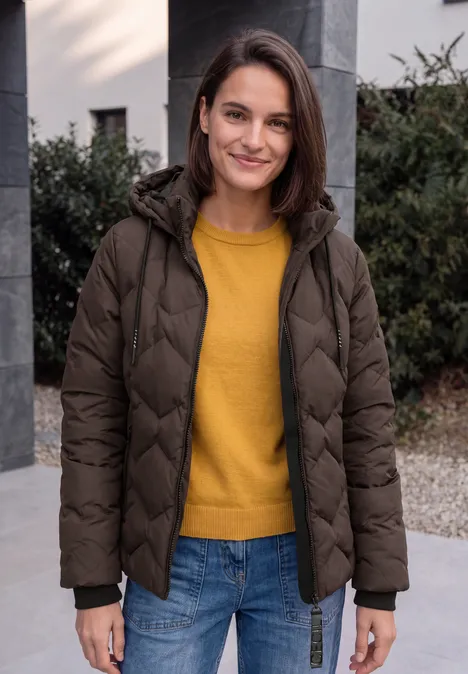 Eine lächelnde Frau in brauner Steppjacke und gelbem Pullover steht draußen.