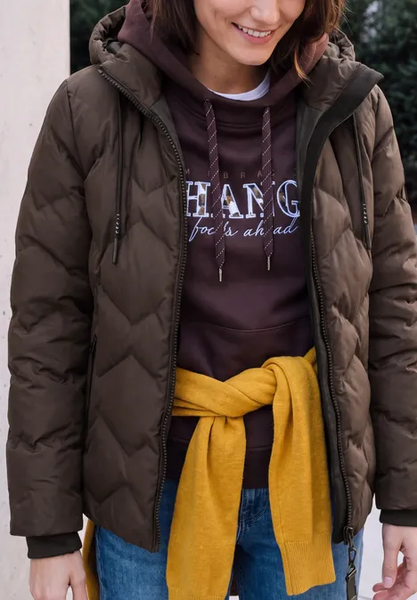 Person mit brauner Steppjacke, violettem Hoodie und gelbem Pullover um die Hüfte, lächelnd.