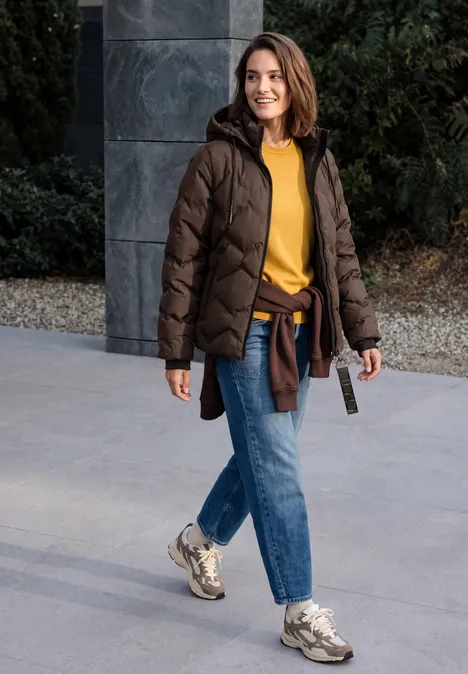 Eine Frau in brauner Steppjacke, gelbem Pullover und Jeans spaziert entspannt im Freien.