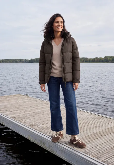 Eine Frau in dunkler Daunenjacke und weiten Jeans steht auf einem Steg am See und lächelt.
